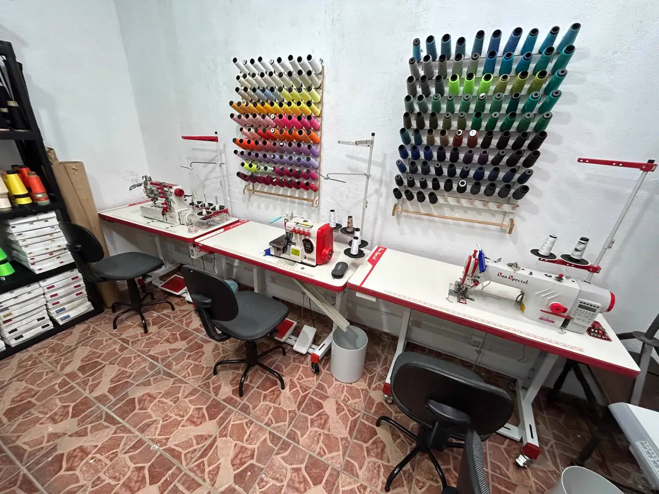 Bancadas com máquinas de costura industriais e paredes de linhas coloridas no ateliê