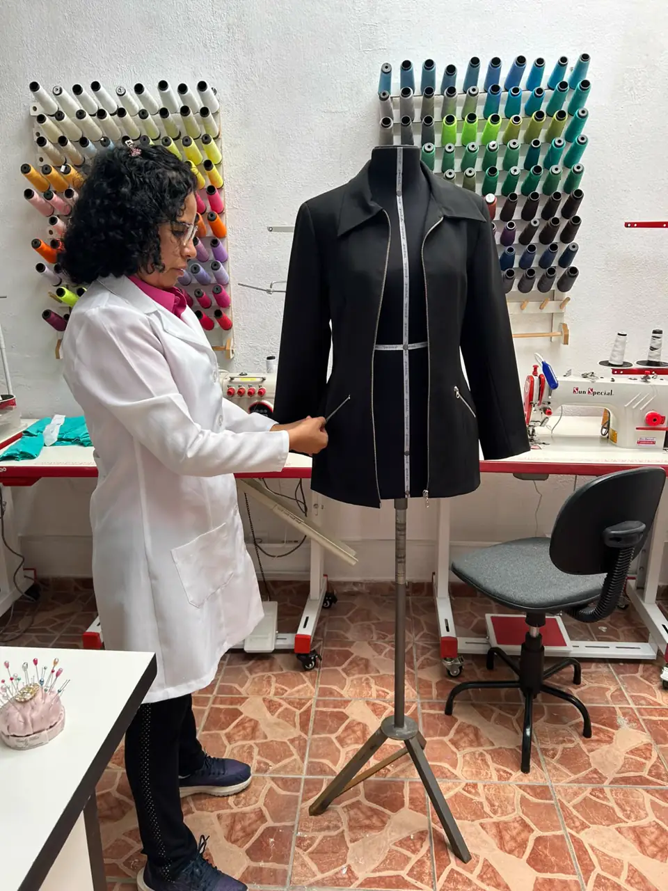Costureira do Luz Escalante Ateliê marcando o ajuste de um blazer preto no manequim, com as paredes de linhas coloridas ao fundo