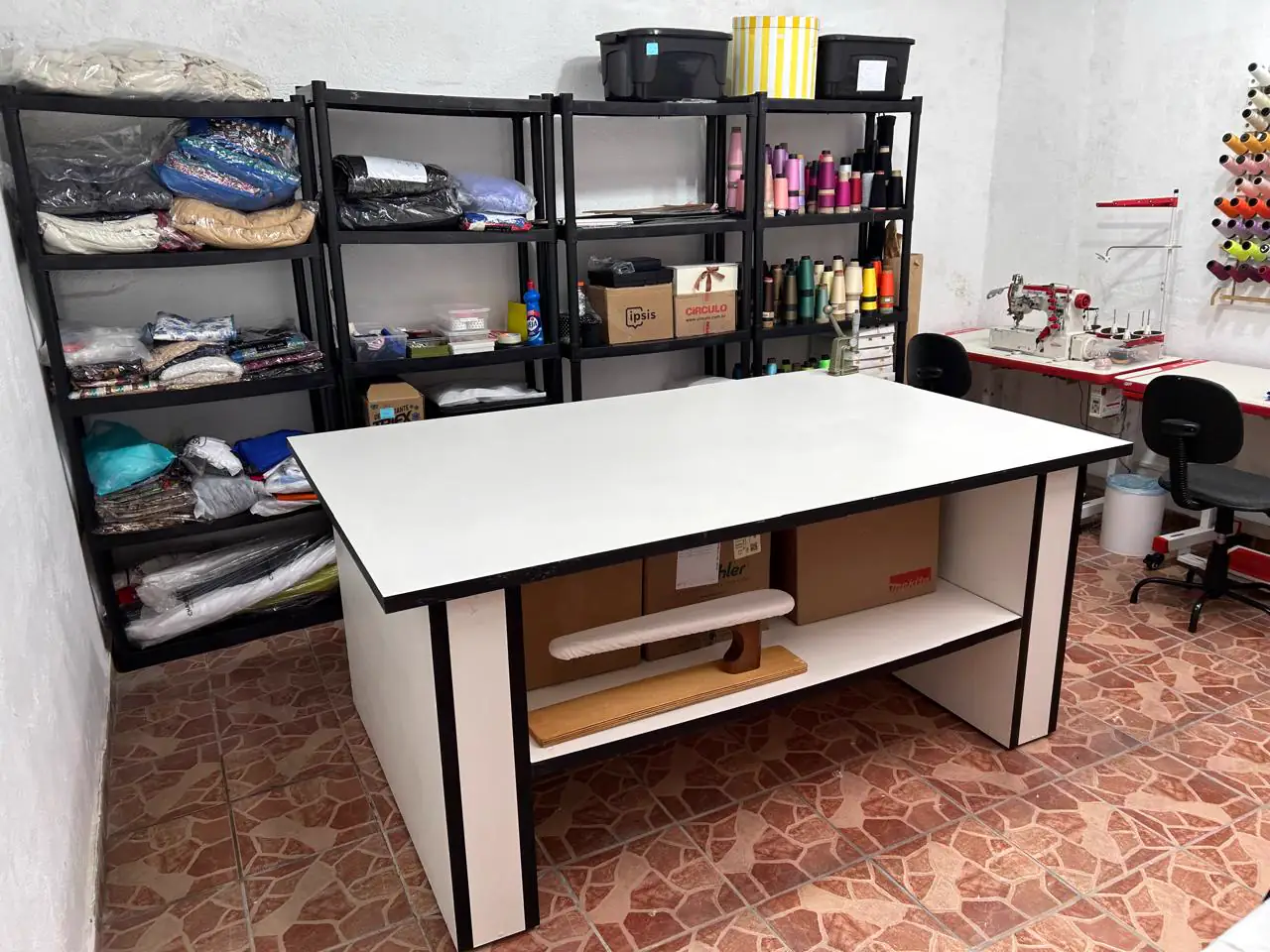 Mesa de corte ampla e estantes com tecidos, linhas e materiais de costura no ateliê